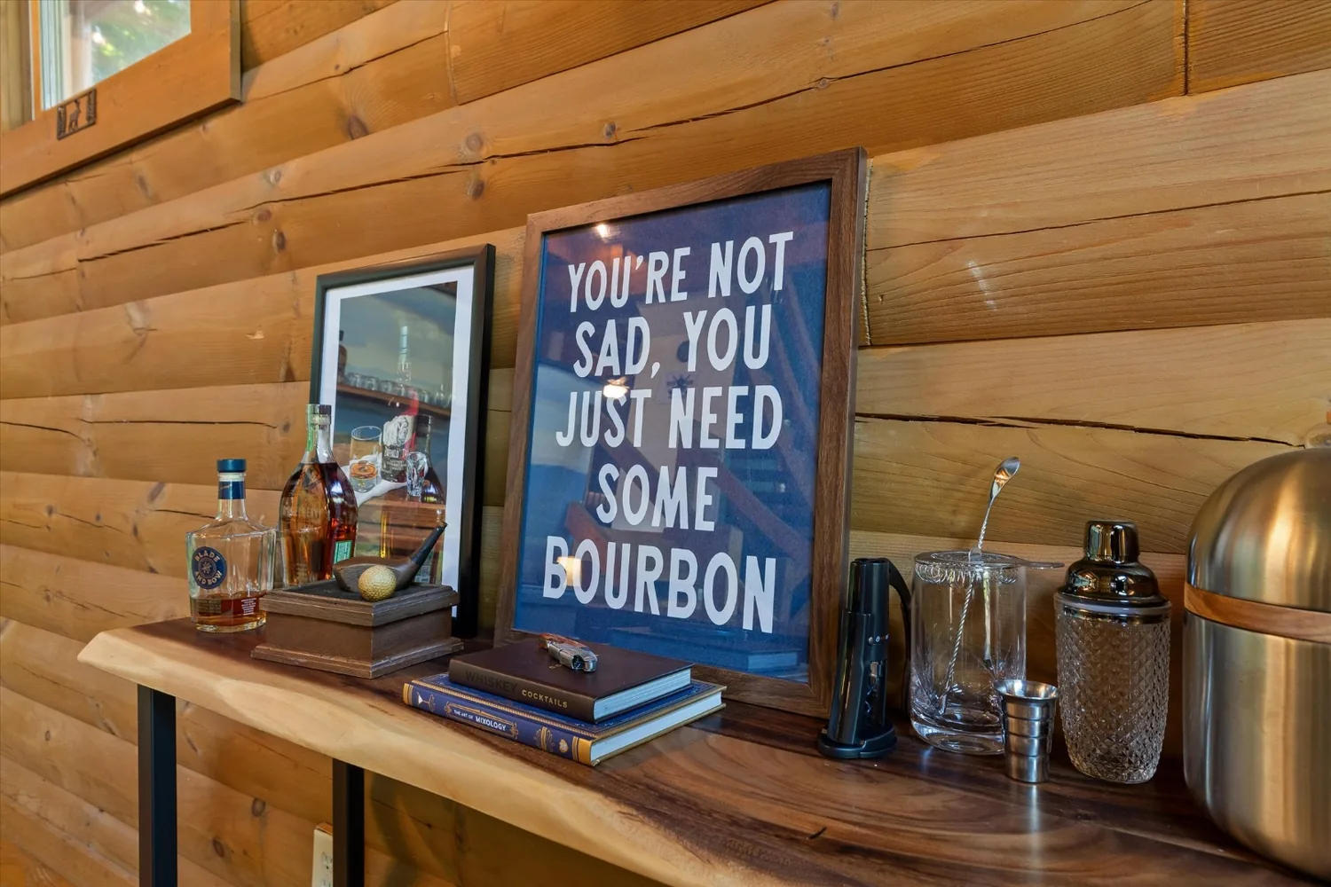 Bar console with bottles, glassware, a shaker and a framed bourbon sign against the log wall